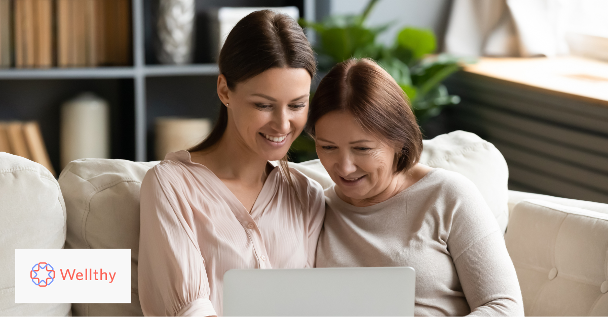 A photo of two woman sitting on a couch looking and smiling at a laptop screen together.